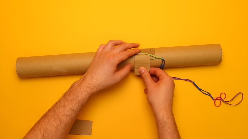 Use your 2 cardboard scraps to attach the rubber bands to the postal tube