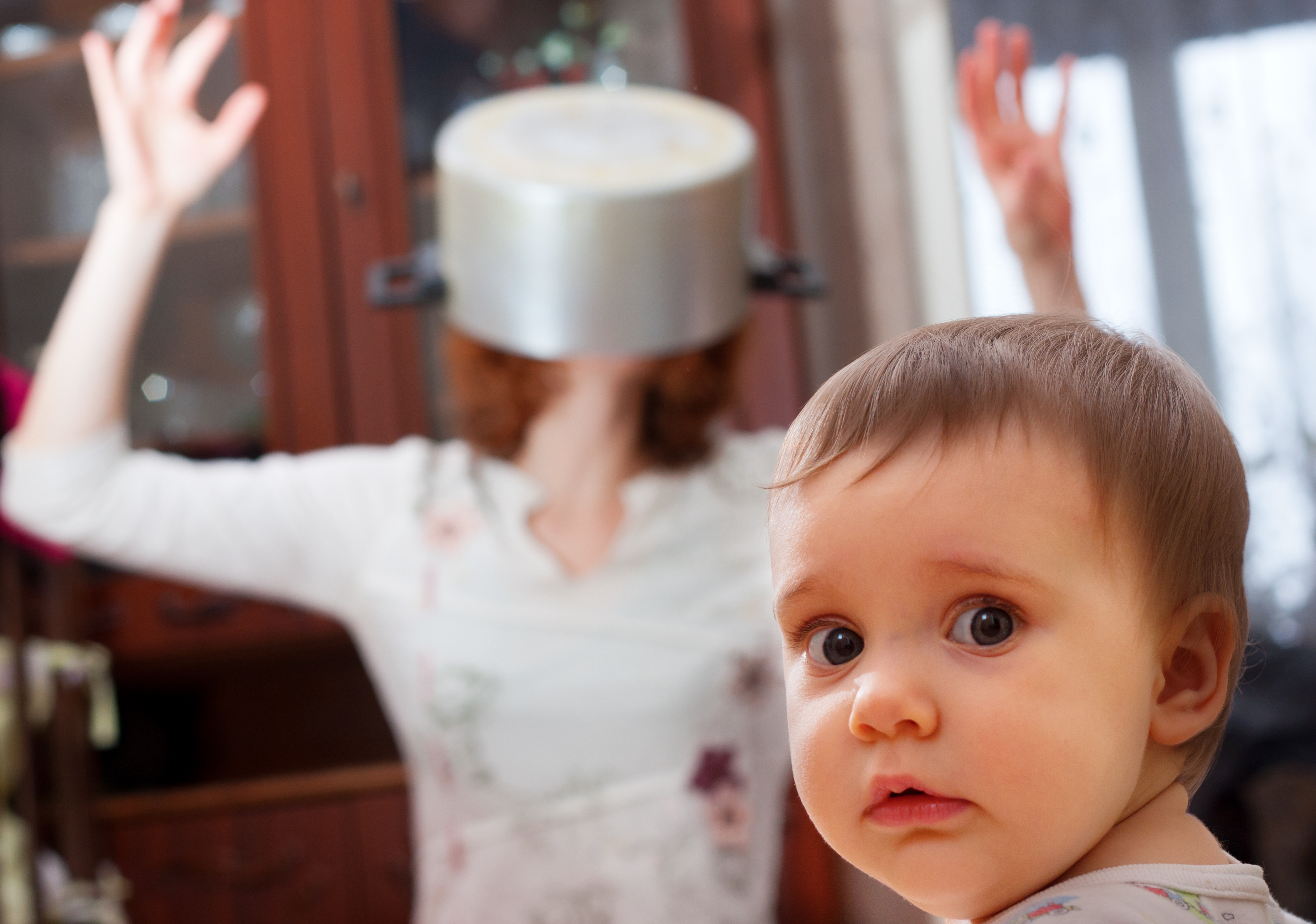 Confused looking baby stares at camera while mother wears saucepan on head in background